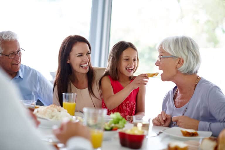 A family gathered around a dining table enjoying dinner together, smiling and interacting. The setting is indoors with various tableware and food items present.